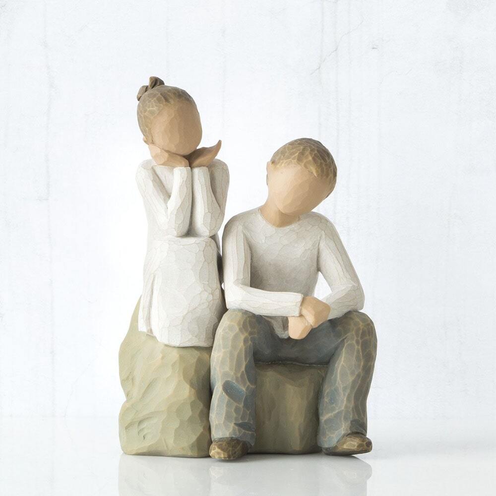 Figure of boy and girl, seated on a gray rock next to each other. Girl in cream dress, boy in cream shirt and blue jeans