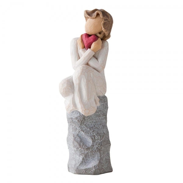 Female figure in cream dress, seated on a gray rock, with knees drawn up to chest, holding red heart up to face. Rock etched with words and symbols of love